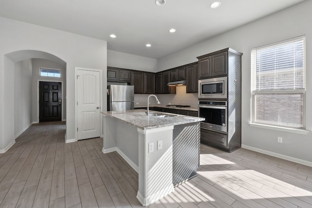 kitchen featuring decorative backsplash, light stone counters, stainless steel appliances, sink, and an island with sink