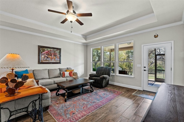 living room with dark wood-style floors, a raised ceiling, ornamental molding, ceiling fan, and baseboards