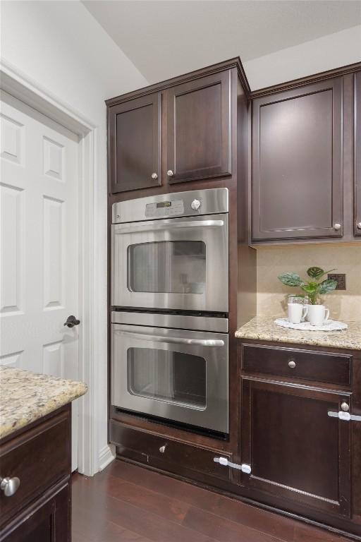 kitchen with decorative backsplash, light stone counters, dark brown cabinetry, double oven, and dark hardwood / wood-style floors