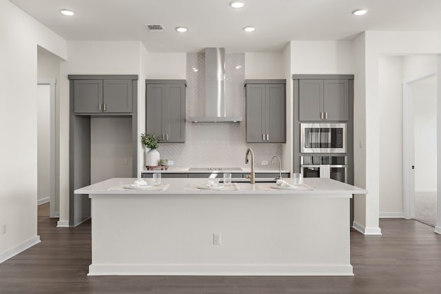 kitchen featuring built in microwave, wall chimney exhaust hood, oven, an island with sink, and gray cabinets