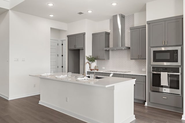 kitchen with gray cabinetry, stainless steel oven, wall chimney range hood, an island with sink, and built in microwave