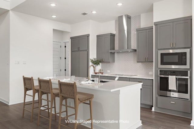 kitchen featuring dark wood-type flooring, wall chimney range hood, a center island with sink, gray cabinets, and oven