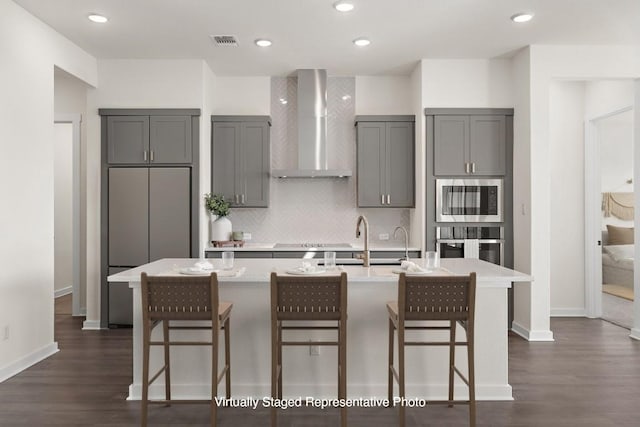 kitchen featuring appliances with stainless steel finishes, a kitchen island with sink, wall chimney exhaust hood, and gray cabinetry