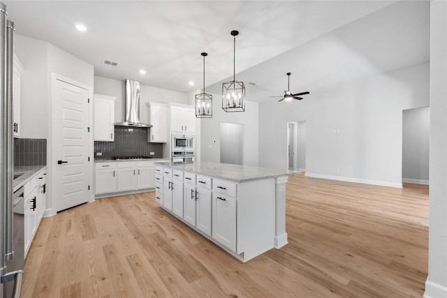 kitchen with ceiling fan, white cabinetry, wall chimney exhaust hood, stainless steel appliances, and a kitchen island