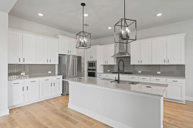 kitchen with white cabinets, an island with sink, hanging light fixtures, and wall chimney range hood