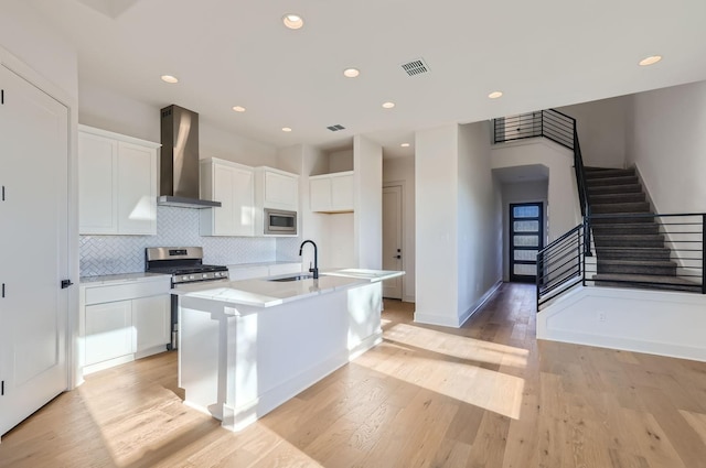 kitchen featuring stainless steel appliances, wall chimney range hood, light wood-type flooring, a kitchen island with sink, and white cabinetry