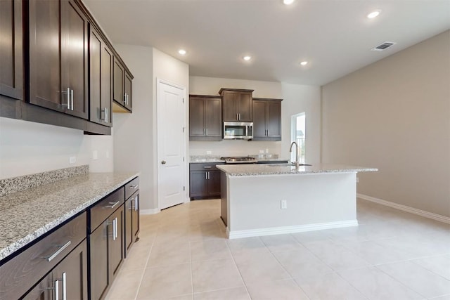 kitchen with light stone countertops, dark brown cabinets, a kitchen island with sink, and sink