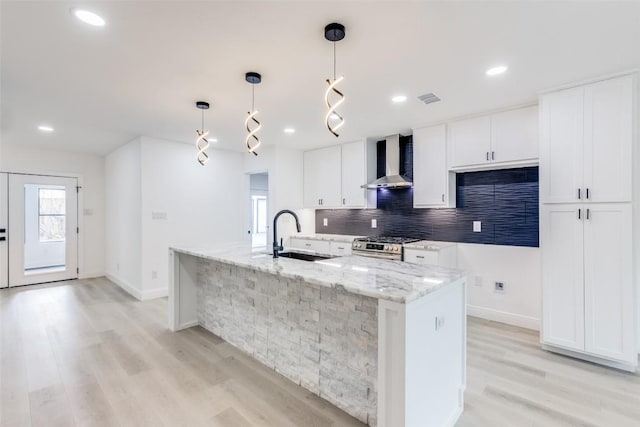 kitchen featuring white cabinetry, sink, hanging light fixtures, wall chimney range hood, and an island with sink