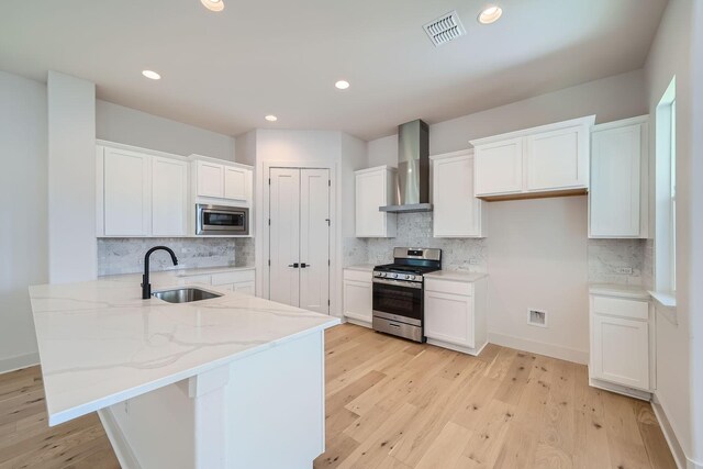 kitchen featuring wall chimney range hood, a kitchen island with sink, white cabinetry, appliances with stainless steel finishes, and sink