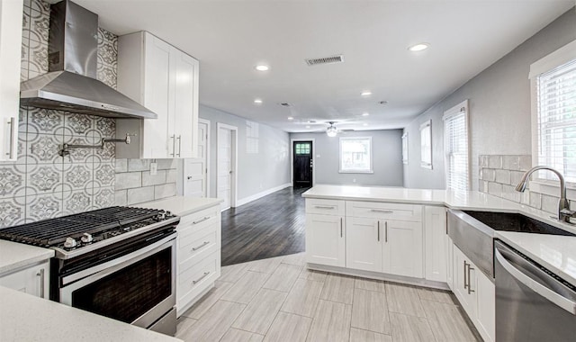 kitchen featuring kitchen peninsula, stainless steel appliances, sink, wall chimney range hood, and white cabinets