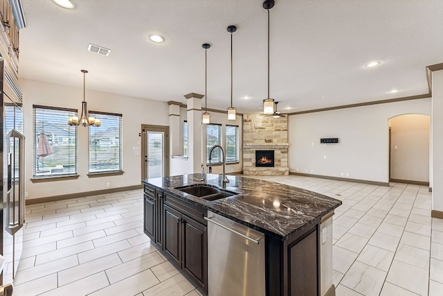 kitchen with dark stone counters, stainless steel appliances, a kitchen island with sink, sink, and a stone fireplace