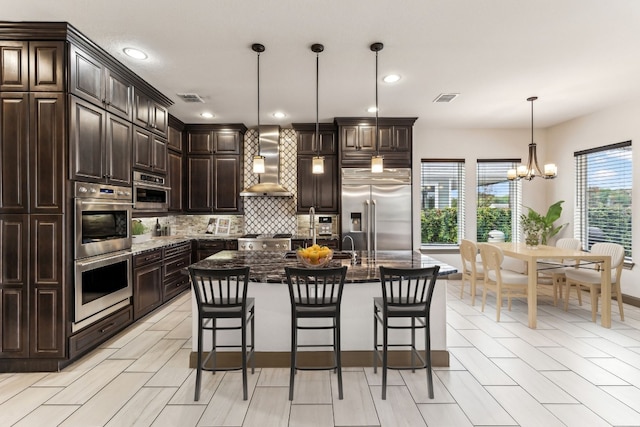 kitchen with a center island with sink, wall chimney range hood, hanging light fixtures, tasteful backsplash, and stainless steel appliances