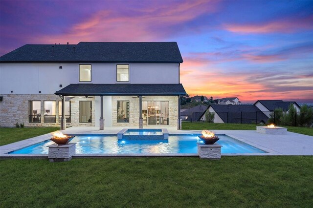 back house at dusk featuring an in ground hot tub, a patio area, a fire pit, and a lawn