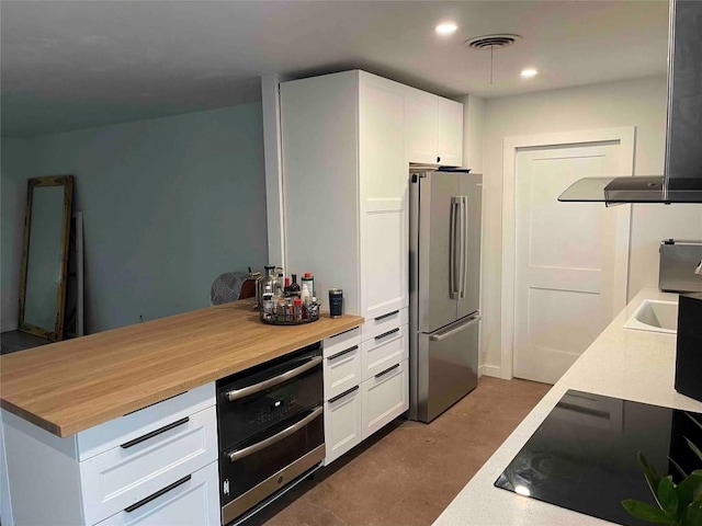 kitchen featuring ventilation hood, black electric cooktop, white cabinets, butcher block countertops, and stainless steel refrigerator