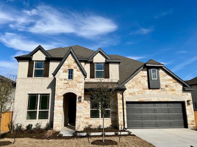 french country home featuring an attached garage, brick siding, fence, driveway, and roof with shingles