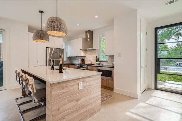 kitchen featuring wall chimney exhaust hood, decorative light fixtures, stainless steel fridge with ice dispenser, white cabinetry, and an island with sink