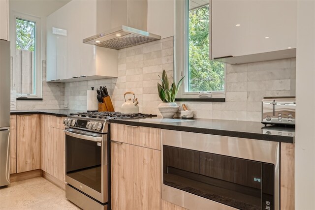 kitchen featuring stainless steel gas stove, light brown cabinets, decorative backsplash, and wall chimney range hood