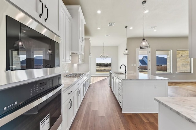 kitchen featuring light stone countertops, sink, white cabinetry, hanging light fixtures, and an island with sink