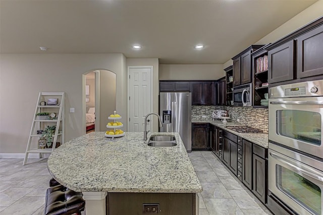 kitchen featuring sink, light stone counters, an island with sink, dark brown cabinets, and appliances with stainless steel finishes