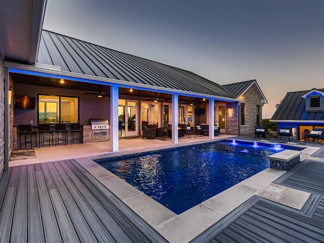 pool at dusk featuring ceiling fan, exterior kitchen, a grill, a hot tub, and exterior bar