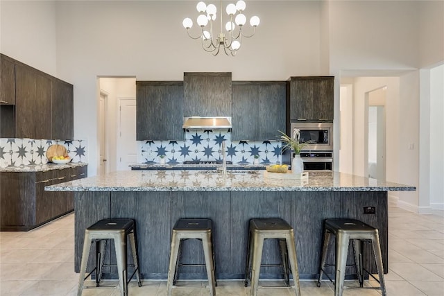 kitchen with a kitchen island with sink, dark brown cabinetry, stainless steel appliances, a high ceiling, and light stone countertops