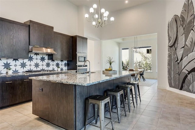 kitchen with modern cabinets, hanging light fixtures, a kitchen island with sink, under cabinet range hood, and a chandelier