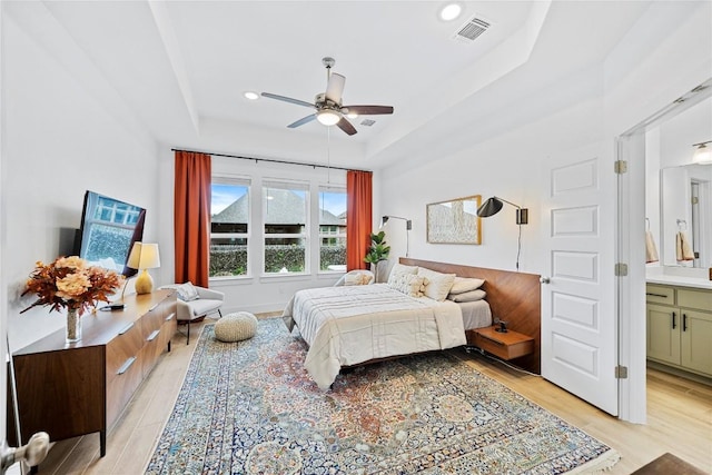 bedroom featuring light wood-type flooring, connected bathroom, a tray ceiling, and ceiling fan