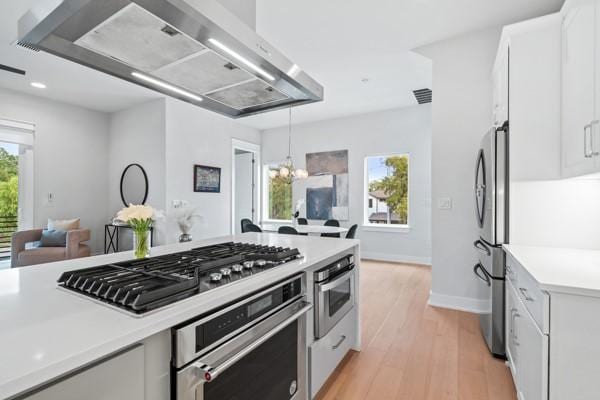 kitchen featuring wall chimney range hood, light hardwood / wood-style flooring, decorative light fixtures, white cabinetry, and stainless steel appliances