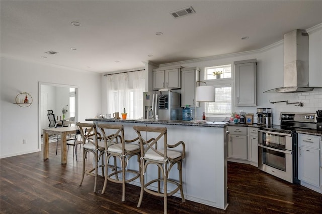 kitchen featuring a healthy amount of sunlight, stainless steel appliances, wall chimney range hood, an island with sink, and gray cabinets