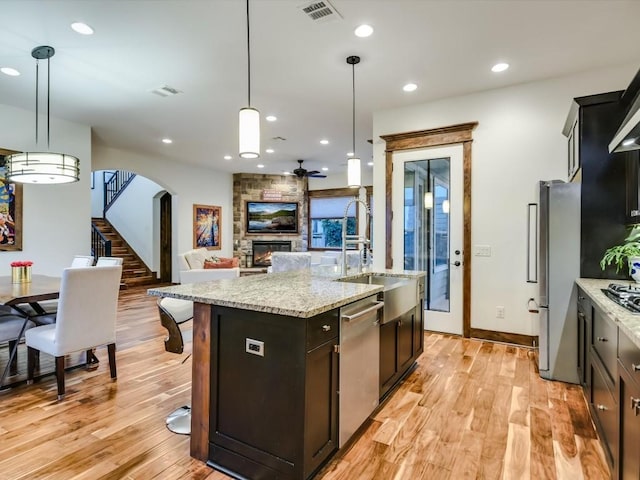 kitchen featuring pendant lighting, a kitchen island with sink, a stone fireplace, ceiling fan, and stainless steel appliances