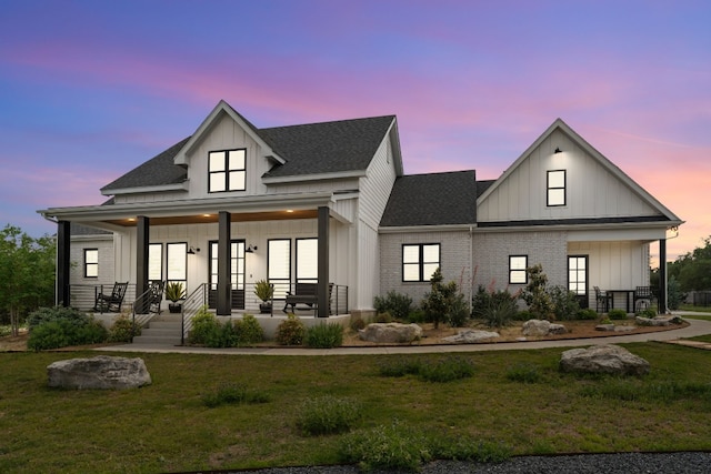 back of property at dusk featuring a shingled roof, a yard, a porch, board and batten siding, and brick siding