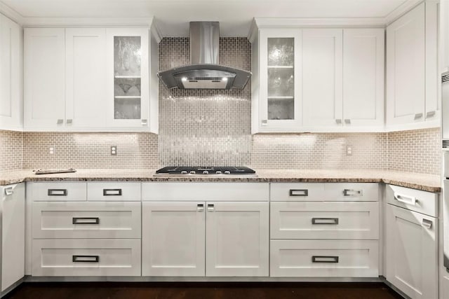 kitchen featuring stainless steel gas stovetop, backsplash, wall chimney exhaust hood, light stone counters, and white cabinetry