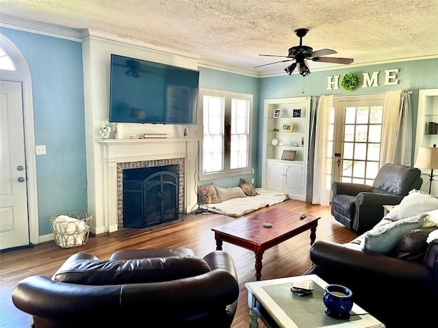 living room with ceiling fan, a brick fireplace, crown molding, hardwood / wood-style floors, and a textured ceiling