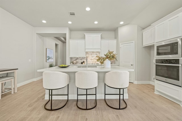 kitchen with stainless steel oven, a kitchen island with sink, white cabinets, light wood-type flooring, and built in microwave