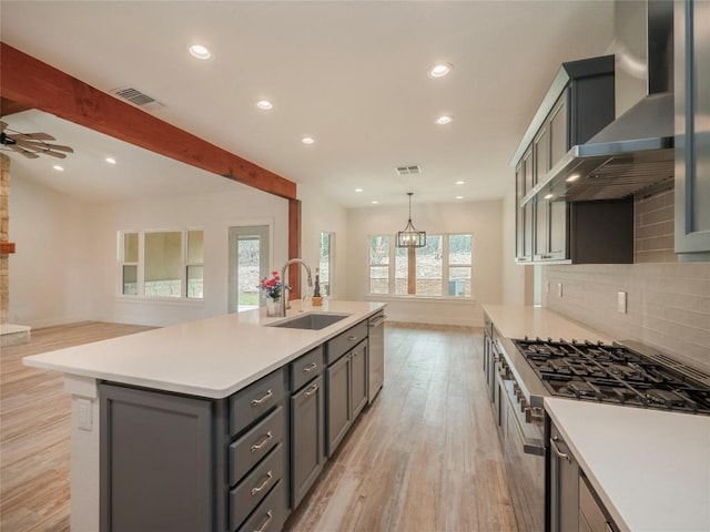 kitchen with sink, wall chimney range hood, beamed ceiling, a kitchen island with sink, and ceiling fan with notable chandelier