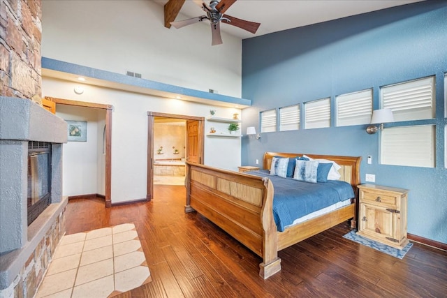 bedroom with a stone fireplace, ensuite bath, ceiling fan, a towering ceiling, and wood-type flooring