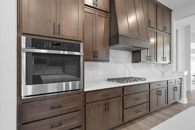 kitchen featuring light stone countertops, backsplash, light hardwood / wood-style floors, appliances with stainless steel finishes, and custom exhaust hood