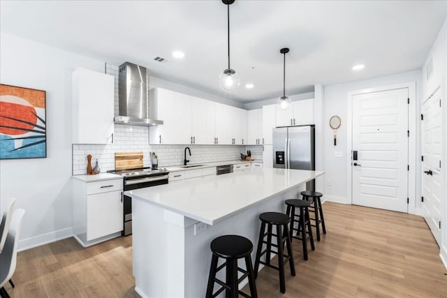 kitchen featuring stainless steel appliances, a kitchen island, wall chimney range hood, white cabinetry, and hanging light fixtures