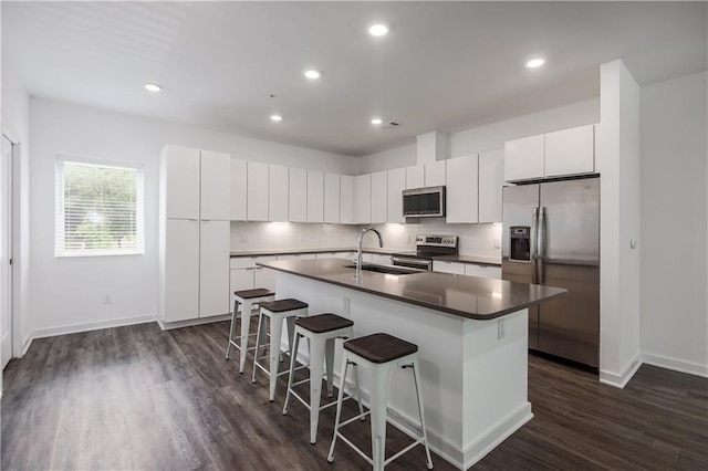 kitchen with a center island with sink, dark countertops, appliances with stainless steel finishes, white cabinets, and a kitchen bar