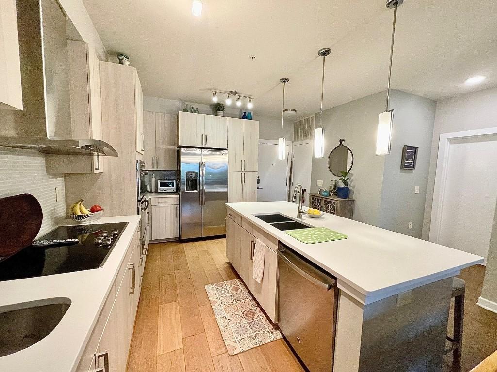 kitchen featuring sink, stainless steel appliances, wall chimney range hood, decorative light fixtures, and a center island with sink