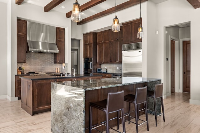 kitchen featuring beam ceiling, wall chimney range hood, dark stone counters, decorative light fixtures, and appliances with stainless steel finishes