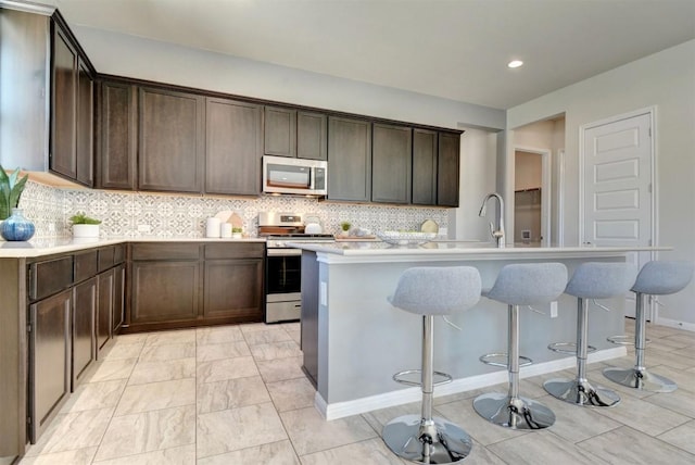 kitchen with appliances with stainless steel finishes, dark brown cabinets, a kitchen island with sink, and backsplash