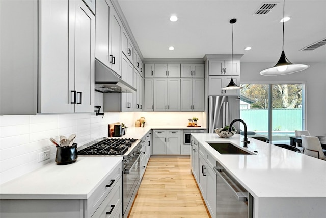 kitchen featuring a kitchen island with sink, sink, hanging light fixtures, appliances with stainless steel finishes, and light hardwood / wood-style floors