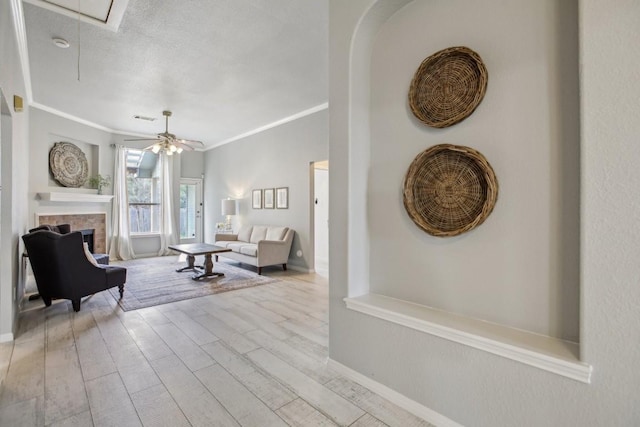 living room with a stone fireplace, ceiling fan, crown molding, a textured ceiling, and light hardwood / wood-style flooring