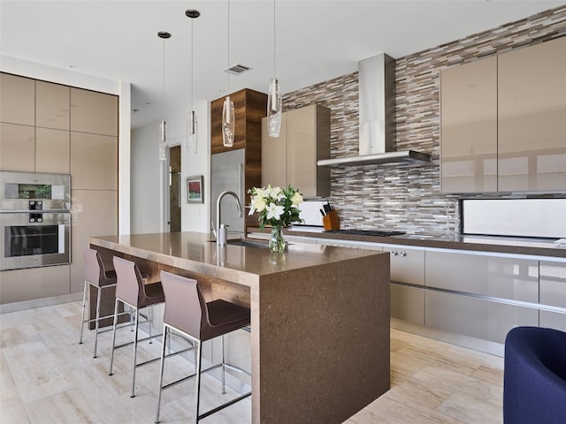 kitchen featuring pendant lighting, a breakfast bar, a kitchen island with sink, wall chimney range hood, and decorative backsplash