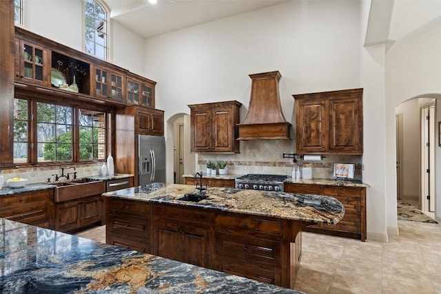 kitchen featuring stainless steel refrigerator with ice dispenser, stove, custom range hood, sink, and stone counters