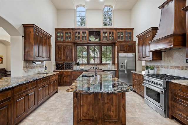 kitchen with stainless steel appliances, premium range hood, dark stone countertops, and a kitchen island with sink