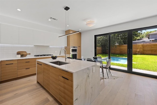 kitchen featuring backsplash, a kitchen island with sink, sink, decorative light fixtures, and white cabinets