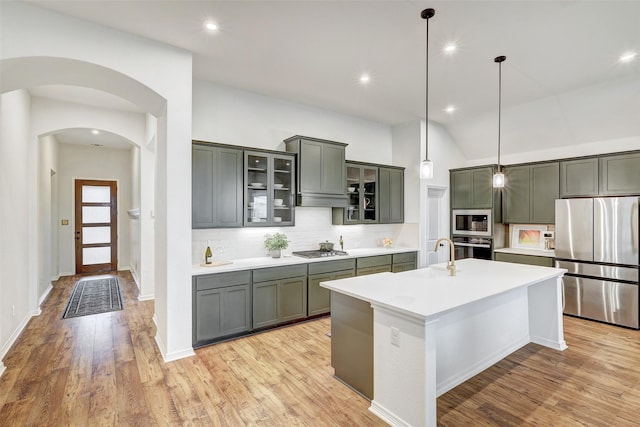 kitchen with light wood-type flooring, pendant lighting, stainless steel appliances, a kitchen island with sink, and decorative backsplash