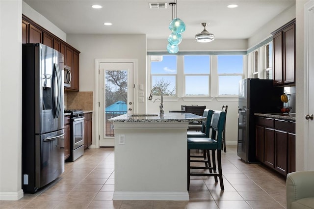 kitchen featuring light stone countertops, hanging light fixtures, stainless steel appliances, a kitchen island with sink, and light tile patterned floors